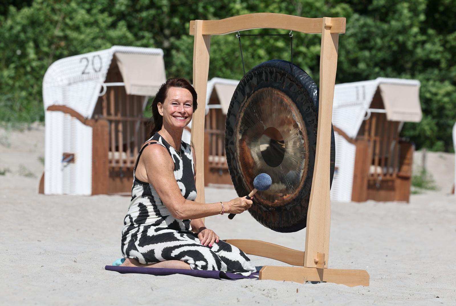Susanne spielt lächelnd einen großen Gong am Strand vor weißen Strandkörben